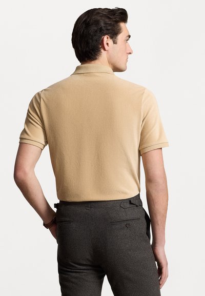 Polo khaki a maniche corte con una texture a coste e polsini piegati, abbinato a pantaloni grigi scuri su misura con tasche posteriori e passanti per la cintura.