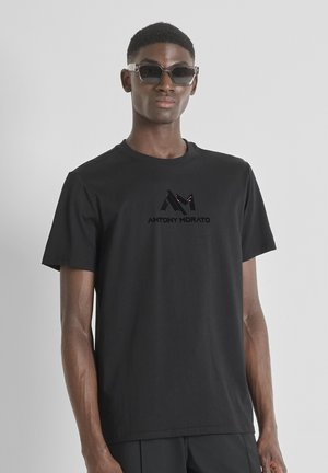 Man wearing black Antony Morato t-shirt and transparent square sunglasses, standing against plain light gray background.