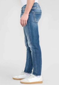 Mies, który ma na sobie dopasowane niebieskie jeansy i białe sneakersy, stoi bokiem na jednolitym tle.