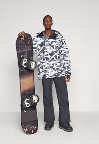 Snowboard με αφηρημένο σχέδιο σε ασπρόμαυρες αποχρώσεις, κρατάει ένα άτομο με jacket με σχέδια, μαύρο παντελόνι και σκούρες μπότες, σε γκρι φόντο.