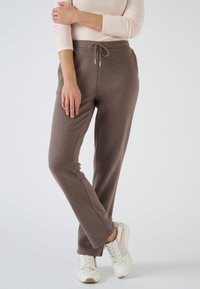 Pantalones de chándal marrones hechos de un tejido suave, con cintura ajustable con cordón, bolsillos laterales y un corte relajado. Llevados con zapatillas blancas y beige.