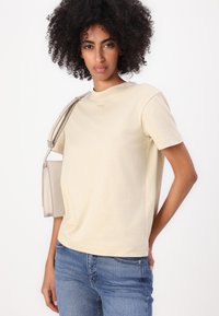 Beige cotton t-shirt with a crew neckline and short sleeves, paired with blue jeans. A small, light-colored bag is slung over one shoulder.