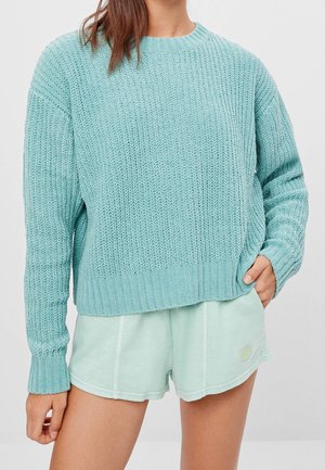 Femme portant un pull en grosse maille bleu clair et un short décontracté vert menthe clair avec une main dans la poche sur un fond uni.