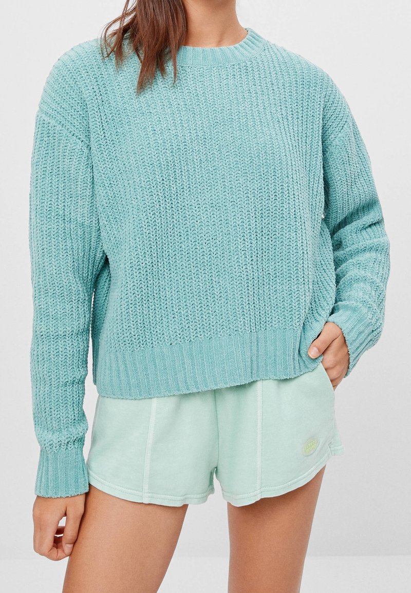 Femme portant un pull en grosse maille bleu clair et un short décontracté vert menthe clair avec une main dans la poche sur un fond uni.