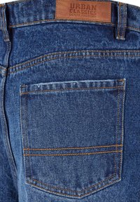 Blå denimjeans med en bakficka, sydd med orange tråd, och en beige läderetikett som står "URBAN CLASSICS DENIM FACTORY."