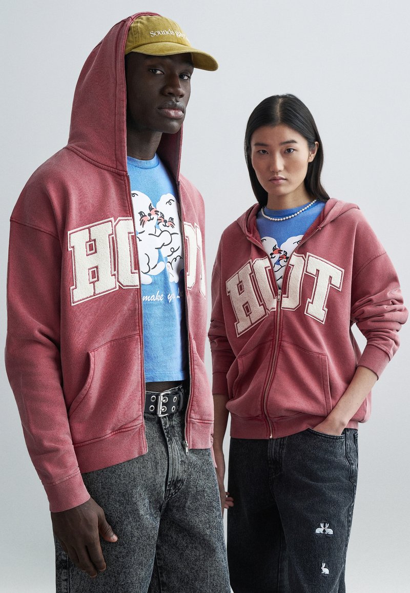 Red hooded sweatshirts with "HOT" text, worn over blue graphic tees. Model wears light denim shorts with rabbit designs. Baseball cap in olive green.