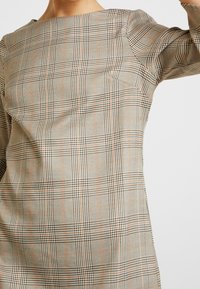 Rutig klänning i beige, bruna och ljusorange nyanser. Har en fyrkantig halsringning och korta ärmar, tillverkad av ett vävt tyg med en slät textur.