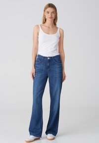 Jean large en denim bleu avec une taille mi-haute, deux poches avant et une fermeture à bouton. Associé à un débardeur ajusté blanc et des baskets.