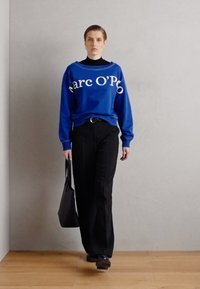 Zils džemperis ar baltu tekstu, melnas bikses un melni apavi. Teksts tuvumā pie apkakles. Modelis nes melnu somu, minimālistisks fons.