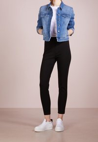 Žena v modré džínové bundě, bílé košili, černých přiléhavých kalhotách a bílých teniskách stojí s rukama v kapsách.