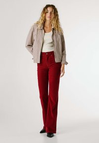 Giovane donna con capelli biondi ricci, che indossa un giubbotto beige a righe, top bianco, pantaloni in velluto a coste rossi e scarpe nere a punta, in piedi.