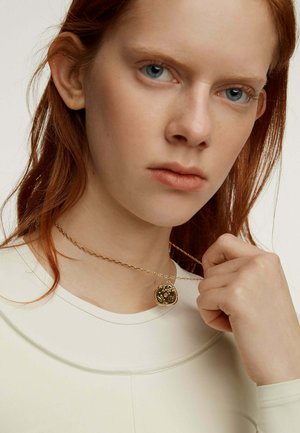 Collar de cadena de oro con un colgante redondo y con patrón, lucido sobre una blusa de color claro, que resalta los intrincados detalles de la joyería.