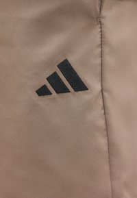 Μαύρο λογότυπο Adidas με τρεις ρίγες κεντημένο σε απαλό ανοιχτό καφέ ύφασμα με ορατή κάθετη ραφή κοντά.