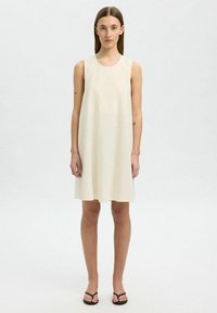 Bezramínková krémová šaty, volný střih, délka ke kolenům, texturovaná tkanina, kulatý výstřih, s jednoduchým, minimalistickým designem. Nosené s černými sandály.