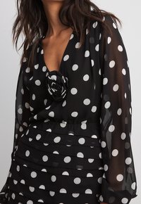 Vestido negro con lunares blancos, con un escote en V profundo, mangas largas transparentes y un adorno de flor de tela en el pecho. Textura rizada en la cintura.