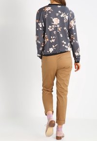 Giubbotto bomber floreale in tessuto grigio scuro con fiori rosa e bianchi, abbinato a pantaloni tapered marrone chiaro e scarpe chiare.