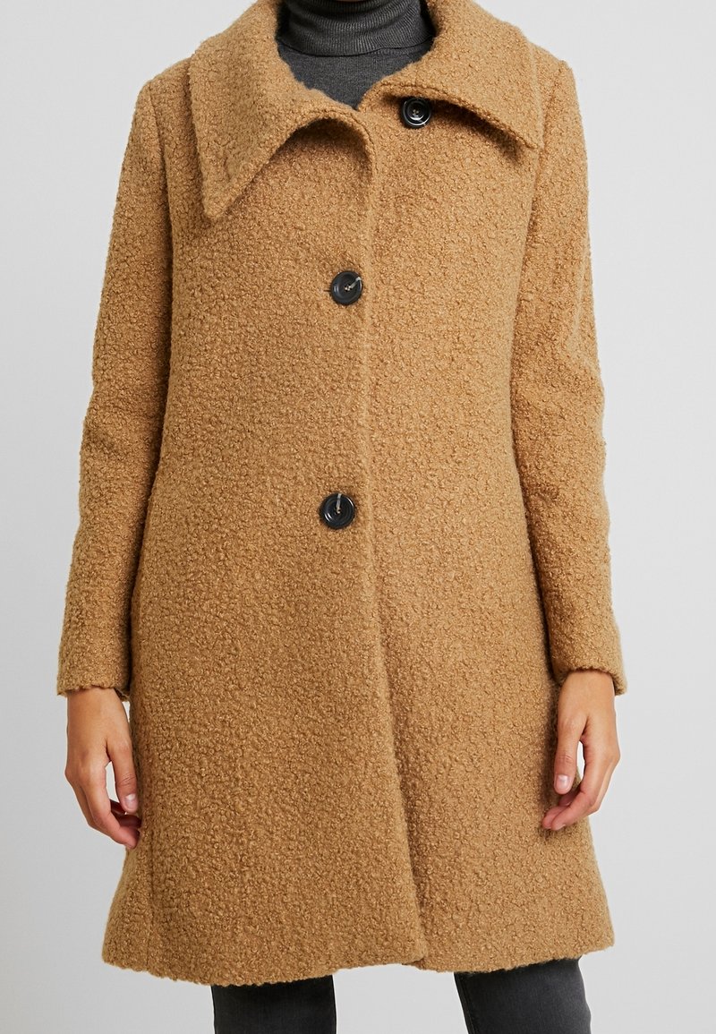 Donna che indossa un cappotto di lana testurizzato color cammello lungo fino al ginocchio, con un ampio colletto e tre bottoni neri, sopra un dolcevita scuro.