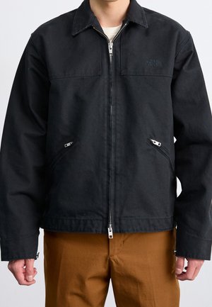 Mężczyzna w czarnej kurtce na zamek z dwoma przednimi kieszeniami na zamek oraz w brązowych spodniach, stojący z rękami swobodnie opuszczonymi wzdłuż ciała.