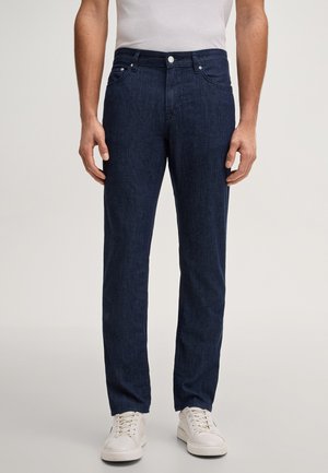 Mężczyzna w ciemnoniebieskich prostych jeansach i białych trampkach, stojący na tle jednolitego jasnego tła.