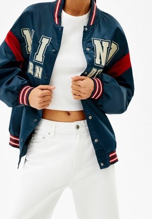 Person iført en marineblå varsityjakke med røde striber, hvid cropped top og højtaljede hvide bukser, mens de holder jakken åbent foran.