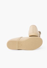 Calzado beige con parte superior de tela suave, puntera redonda, correa ajustable y suela de goma texturizada con un agarre estampado.