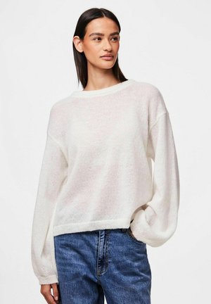 Biały, oversize'owy sweter wykonany na szydełku, z okrągłym dekoltem, długimi rękawami i prążkowanymi mankietami. W połączeniu z niebieskimi dżinsami. Miękka faktura.