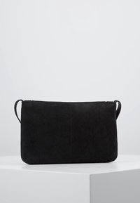 Černá semišová obdélníková kabelka s zápěstním popruhem na šedobílém podstavci na světle šedém pozadí.