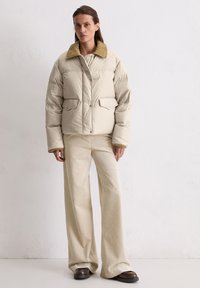 Jachetă puffer bej cu guler galben, închidere cu nasturi și două buzunare frontale, purtată cu pantaloni široi crem cu nervuri și încălțări închise la culoare.