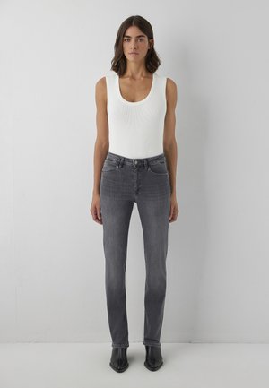 Woman with shoulder-length dark hair wearing a white sleeveless top, gray high-waisted jeans, and black pointed boots standing against a gray wall.