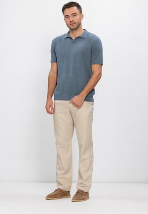 Bărbat purtând tricou polo cu mâneci scurte albastru, pantaloni bej și pantofi maro, stând în picioare pe un fundal alb simplu.