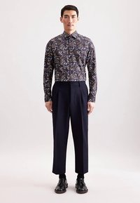 Camicia a maniche lunghe blu navy con stampa floreale e colletto con bottoni, abbinata a pantaloni neri a vita alta e scarpe nere, in piedi.