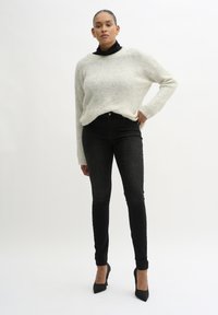 Svijetlosivi, labavo pleteni pulover s uzorkom turtleneck, uparen s hlačama od crnog tankog trapera visokog struka i crnim cipelama na visoku petu.