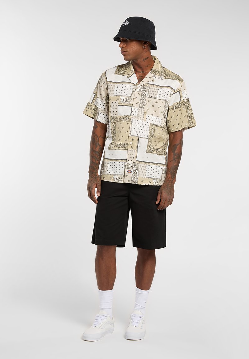 Chemise à manches courtes beige avec divers motifs imprimés ; associée à un short noir. Accessoires : un bob noir et des baskets blanches.