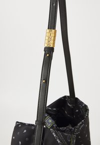 Melna ādas rokassomu siksna ar zeltītām burtiem, kas veido vārdu "PINKO", un zem šīs siksnas ir melna rakstaina auduma soma.