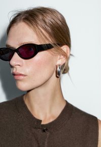 Des lunettes de soleil noires en forme d'œil de chat avec des verres violet profond, accompagnées d'un top en tricot marron avec un col rond et un détail de bouton unique.