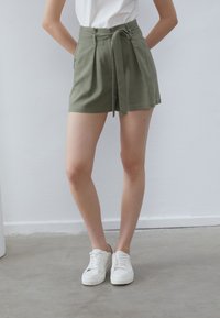 Pantaloni scurți verzi din in, cu talie înaltă, pliseuri și o curea legată. Asortați cu adidași albi pe un podea neutră.