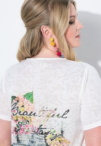 Camiseta blanca semi-translúcida con un estampado floral y la palabra "Hermosa" en una fuente cursiva. Pendientes texturizados y coloridos añaden acentos vibrantes.