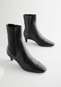 Bottines en cuir noir avec un bout pointu et un petit talon. Elles comportent une fermeture éclair et des panneaux latéraux en tissu pour un ajustement parfait.