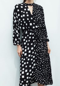 Schwarzes Kleid mit weißen Quadratmuster, Wickeldesign, langen Ärmeln und einem Bindegürtel. Leichter Stoff mit leicht strukturierter Oberfläche.