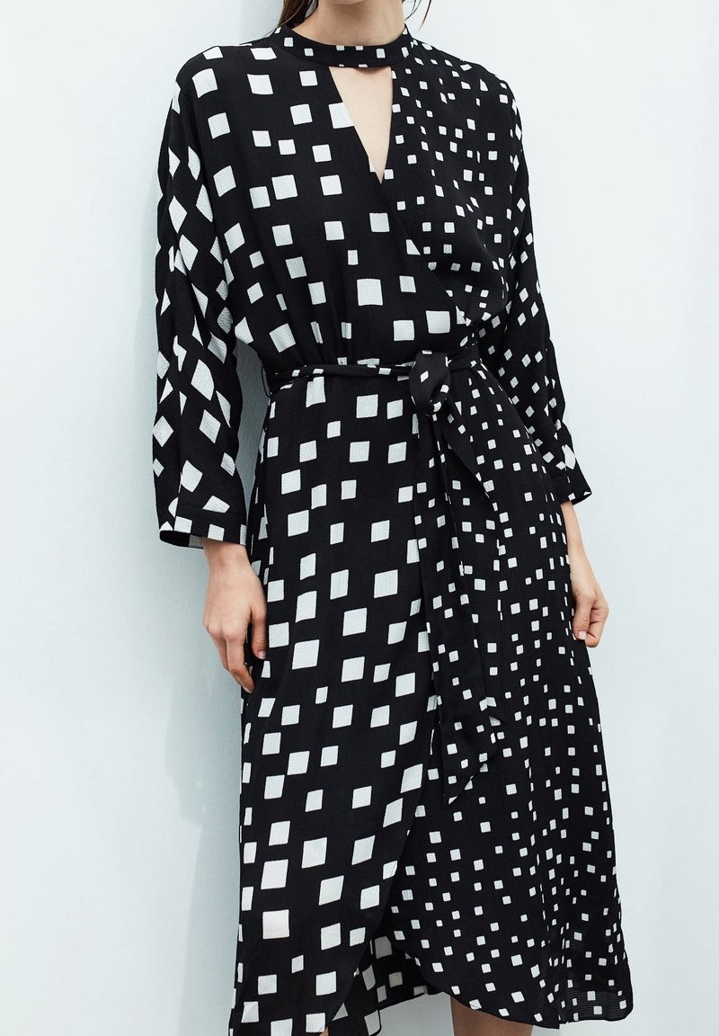Schwarzes Kleid mit weißen Quadratmuster, Wickeldesign, langen Ärmeln und einem Bindegürtel. Leichter Stoff mit leicht strukturierter Oberfläche.