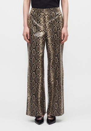 Personne portant un pantalon large imprimé peau de serpent marron et noir avec des chaussures noires pointues, debout sur un fond blanc.