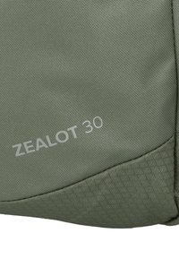 Primo piano di un tessuto verde con un motivo testurizzato e "ZEALOT 30" stampato in grigio chiaro sulla superficie.