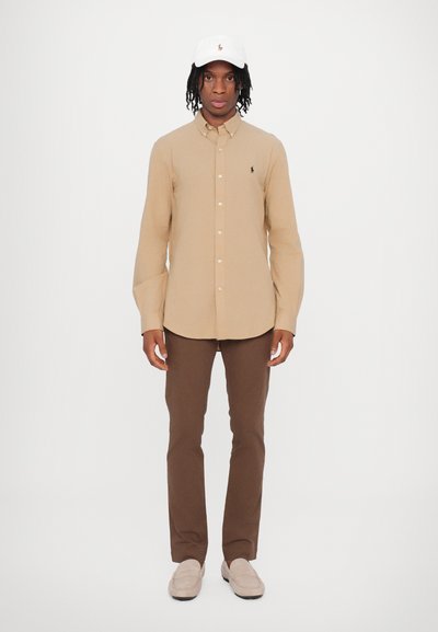 Camisa bege abotoada, calças castanhas, boné branco com logótipo, sapatos claros. A camisa tem um colarinho e um logótipo subtil no peito.