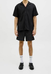 Uomo che indossa una camicia nera a maniche corte con bottoni e pantaloni corti neri abbinati, con calzini bianchi e scarpe nere, in piedi contro uno sfondo neutro.
