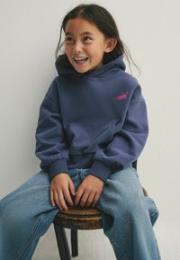 Sudadera con capucha azul marina y logo rosa, bolsillo frontal y puños acanalados, combinada con unos vaqueros de pierna ancha azul claro. Sentada en un taburete de madera.
