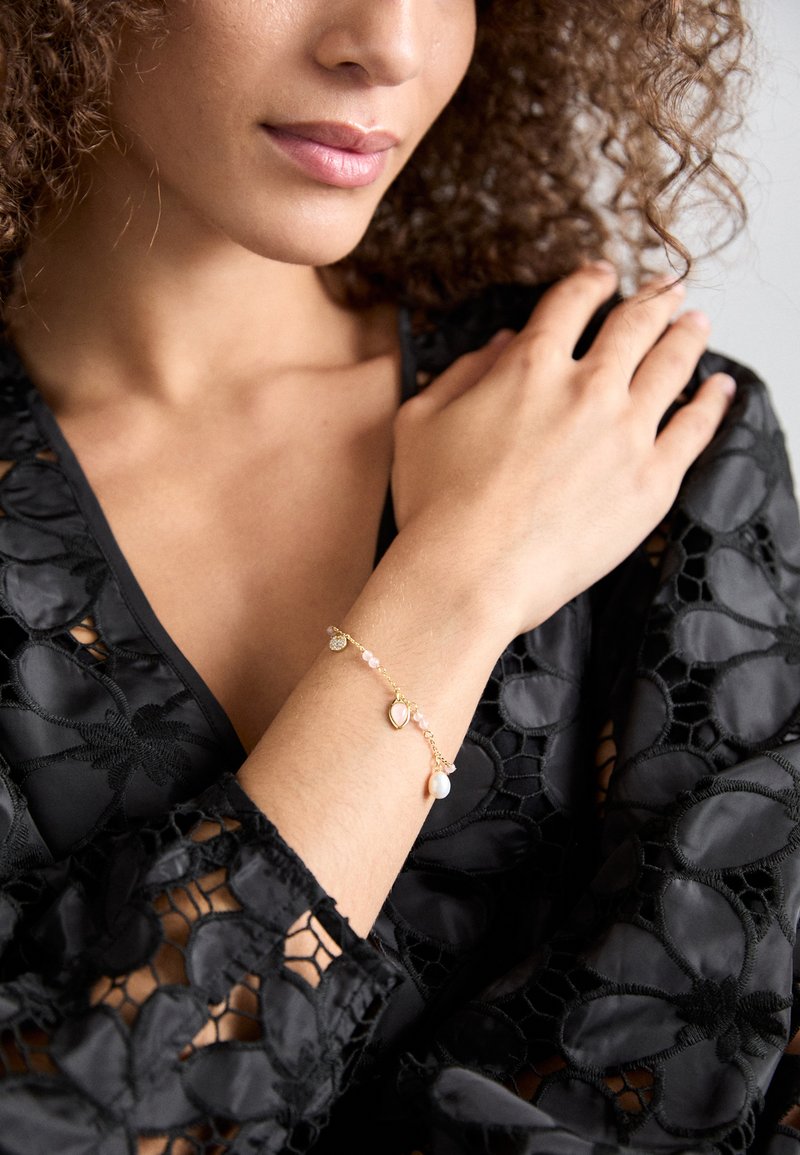 Zlatý náramek s růžovými korálky, kapkovým přívěskem a perlou, nošený na ruce opřené o černou, vyšívanou látku.