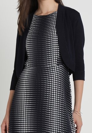 Femme portant une robe à pois noir et blanc avec un cardigan noir court et des bracelets argentés à son poignet droit.
