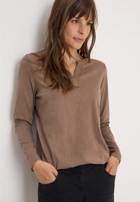 Haut marron à manches longues avec un plastron partiel de boutons, texture douce et coupe décontractée, associé à un pantalon noir et une pose décontractée.