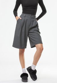 Pantaloncini grigi a righe sottili con vita alta, pieghe e chiusura con bottone. Il modello indossa scarpe nere pelose e calzini bianchi alla caviglia.