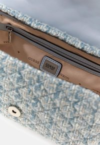 Sininen tweed-käsilaukku, jossa on hopeiset yksityiskohdat, vetoketjulla varustettu tasku ja beige sisustus, jossa on brändin logo ja painettu kuvio.